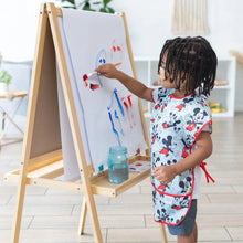 Load image into Gallery viewer, A child in a Bumkins Short-Sleeved Smock: Mickey Mouse Classic paints vibrant strokes at an easel on wooden floors in a bright room.
