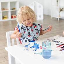 Load image into Gallery viewer, A child with curly blonde hair paints at a white table in a bright room, wearing a Bumkins Short-Sleeved Smock: Mickey Mouse Classic.
