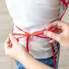 Load image into Gallery viewer, Close-up of hands tying the adjustable ribbon closure of a Bumkins Short-Sleeved Smock: Mickey Mouse Classic around a child's waist.
