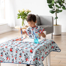 Load image into Gallery viewer, A child paints at a table in a Bumkins Short-Sleeved Smock: Mickey Mouse Classic, amidst a blue water jar, art supplies, and a potted plant.
