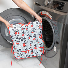 Load image into Gallery viewer, A person holds a Bumkins Short-Sleeved Smock: Mickey Mouse Classic in front of an open front-loading washing machine.
