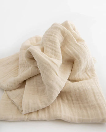 A beige cotton muslin swaddle from the Little Unicorn Cotton Swaddle Set, Woof, laid out on a white surface.