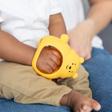 Load image into Gallery viewer, A child holds a Bumkins Silicone Un-Droppable Mitt Teether: Winnie the Pooh, while sitting on an adult’s lap.
