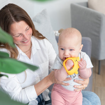 A smiling woman holds a baby with a Bumkins Silicone Un-Droppable Mitt Teether: Winnie the Pooh in a cozy room, plant in view.