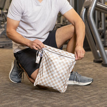Load image into Gallery viewer, A man kneels on a gym floor, unzipping a Roamio by Bumkins Wet Bag: Sand Check.
