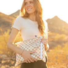 Load image into Gallery viewer, A woman carries a Roamio by Bumkins Wet Bag: Sand Check in golden sunlight with mountains in the background.
