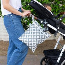 Load image into Gallery viewer, A person wearing jeans and a white shirt pushes a child in a stroller outdoors, with a Bumkins Wet Bag: Charcoal Check attached to the handle.
