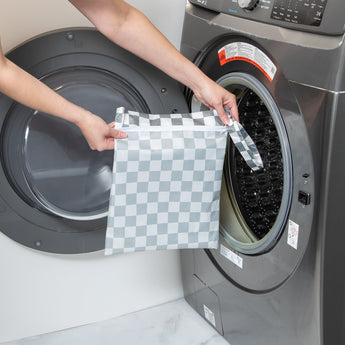 A person holds Bumkins Wet Bag: Charcoal Check in front of an open front-loading washing machine.