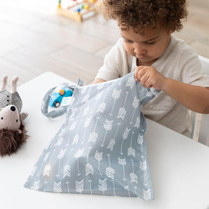 A child at a white table opens a Bumkins Wet Bag: Arrow, with a toy car and stuffed animal nearby.
