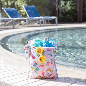 Roamio by Bumkins Wet Bag: Boots holds yellow sunglasses, a towel, water bottle, swimwear, & goggles poolside; empty lounge chairs behind.