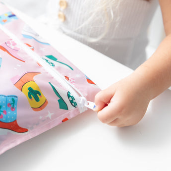 A child's hand unzips a Roamio by Bumkins Wet Bag: Boots, crafted from colorful and waterproof fabric and designed for reuse.