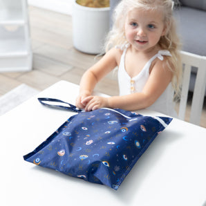 A child with blonde hair smiles at a table, holding Bumkins Wet Bag: Evil Eye.