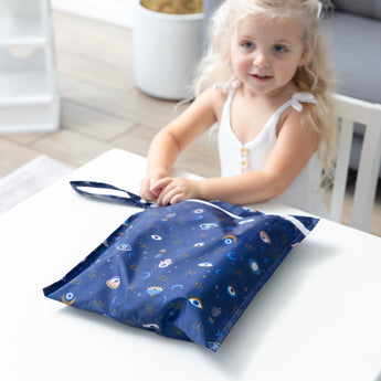 A child with blonde hair smiles at a table, holding Bumkins Wet Bag: Evil Eye.