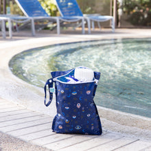 Load image into Gallery viewer, Bumkins Wet Bag: Evil Eye holds a towel and swimwear poolside with empty blue lounge chairs behind.
