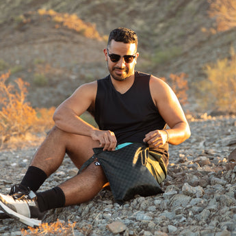 A man wearing sunglasses sits on rocky ground in golden sunlight, reaching into a Roamio by Bumkins Wet Bag: Rich Black.