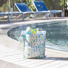 Load image into Gallery viewer, Bumkins Wet Bag: Cacti sits poolside, holding swimwear and a water bottle with empty lounge chairs in the background.
