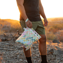 Load image into Gallery viewer, A person wearing green shorts and a black shirt stands on rocky ground at sunset, holding a Roamio by Bumkins Wet Bag: Motley Check.
