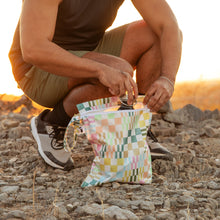 Load image into Gallery viewer, A man crouches on rocky ground in golden sunlight, placing a water bottle into a Roamio by Bumkins Wet Bag: Motley Check.
