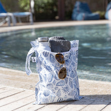 Load image into Gallery viewer, Roamio by Bumkins Wet Bag: Shells holds sunglasses, a towel, and water bottle poolside.
