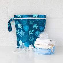 Load image into Gallery viewer, Bumkins Wet Bag: Blue Tropic sits on a white table against a white brick wall, with cloth diapers and breast pump parts in front.
