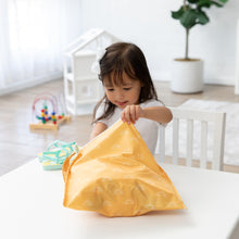 Load image into Gallery viewer, A young girl at a white table opens a Bumkins Wet Bag: Sunshine, with a playhouse and toys brightening the airy room.
