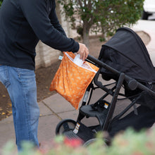 Load image into Gallery viewer, A person places a bottle into a Bumkins Wet Bag: Grounded attached to a stroller handle on a paved path with greenery.
