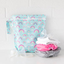 Load image into Gallery viewer, Bumkins Wet Bag: Rainbows sits on a white counter against a white brick wall with cloth diapers and breast pump parts in front.
