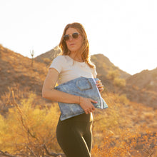 Load image into Gallery viewer, A woman in sunglasses holds a Bumkins Wet Bag: Vintage Denim in golden sunlight with mountains behind.
