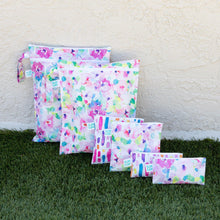 Load image into Gallery viewer, Bumkins Wet Bag: Watercolor sits among other similarly printed bags on grass against a textured beige wall.
