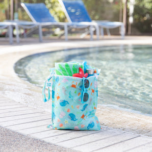 Bumkins Wet Bag: Ocean Life holds sunglasses, a towel, and swim gear poolside with empty lounge chairs in the background.