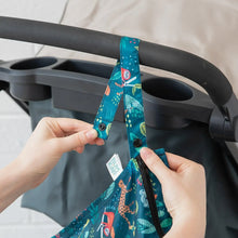 Load image into Gallery viewer, Close-up of hands attaching a Bumkins Wet Bag: Jungle to a stroller handle using the snap button on the strap.
