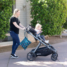 Load image into Gallery viewer, A woman pushes a stroller with a toddler inside, a Bumkins Wet Bag: Jungle hanging from the handle; trees and a building in the background.

