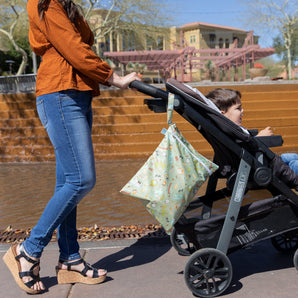 A woman pushes a child in stroller near a fountain, with a Bumkins Wet Bag: Camp Gear hanging from the handle under bright sun.