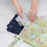 Hands placing a gray shirt and denim jeans into Bumkins Wet Bag: Camp Gear on a marble surface.
