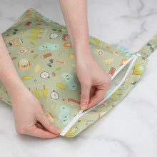 Load image into Gallery viewer, Hands zipping a Bumkins Wet Bag: Camp Gear, featuring trees and tents on waterproof fabric, on a marble surface.
