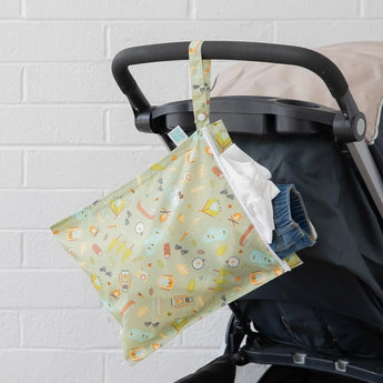 A stroller in front of a white wall with a Bumkins Wet Bag: Camp Gear hanging from the handle, filled with a white shirt and denim shorts.