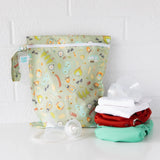 Cloth diapers and breast pump parts sit on a white table in front of a Bumkins Wet Bag: Camp Gear against a white brick wall.