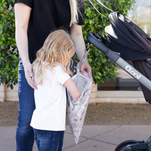 Load image into Gallery viewer, A child peeks into a Bumkins Wet Bag: Floral attached to a stroller handle, with an adult nearby outdoors.

