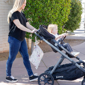 A woman walks with a toddler in a stroller on a sidewalk near greenery, with Bumkins Wet Bag: Floral hanging from the handle.
