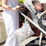A person pushes a stroller with a baby; a Bumkins Wet Bag: Mickey & Friends Varsity hangs on the handle.