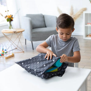 A child in a gray shirt opens a Bumkins Wet Bag: Mickey Mouse Icon Black + White at a white table in a living room with toys and flowers.