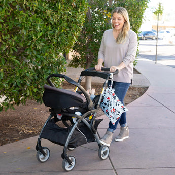 A smiling woman in jeans pushes a stroller with a Bumkins Wet Bag: Mickey Mouse Classic attached to the handle on a sidewalk past greenery.