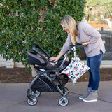 A woman with long hair leans over a stroller on a sidewalk, with a Bumkins Wet Bag: Mickey Mouse Classic attached to the handle.