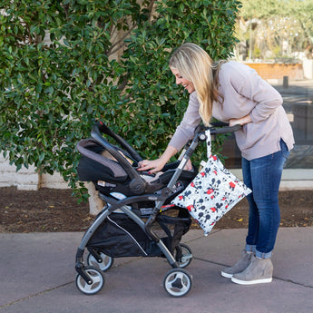 A woman with long hair leans over a stroller on a sidewalk, with a Bumkins Wet Bag: Mickey Mouse Classic attached to the handle.