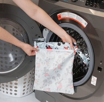 A person holds Bumkins Wet Bag: Mickey Mouse Classic in front of an open front-loading washing machine.
