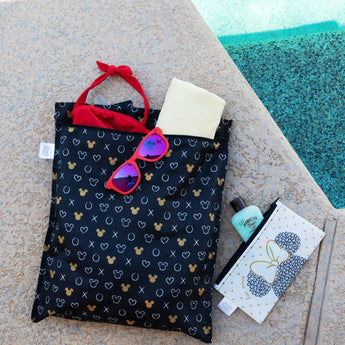 Bumkins Wet Bag: Minnie Mouse Icon Black + White sits by a pool, with sunglasses, a towel, and a swimsuit peeking out; another bag is nearby.