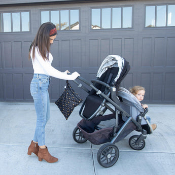 A woman pushes a child in a stroller by a garage, with a Bumkins Wet Bag: Minnie Mouse Icon Black + White hanging from the handle.