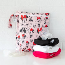 Load image into Gallery viewer, Bumkins Wet Bag: Minnie Mouse Classic sits on a white table against a white brick wall with cloth diapers and breast pump parts in front.
