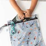 A child slips a book into a Bumkins Wet Bag: No Tricks, Just Treats on a white table, with blue sunglasses nearby.