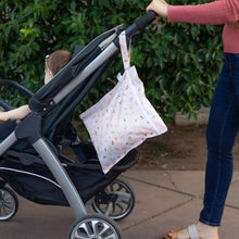 Load image into Gallery viewer, A person strolls with a baby in a stroller and Bumkins Wet Bag: Princess Magic hanging from the handle along a sidewalk lined with greenery.
